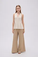Blusa de algodón color beige con escote en V, cuello halter y cierre frontal con botones. Presenta un diseño ajustado en el torso con pinzas y pliegues en la parte inferior que generan una caída ligeramente acampanada.