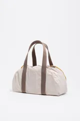 Mini bolso de mano Bimba y Lola con estructura blanda de nylon en color beige. Presenta asas de cinta en tono marrón, cierre mediante cordones laterales amarillos con detalles metálicos y logo metálico frontal.