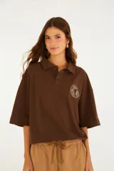 Remera tipo polo marrón, de corte oversize, con cuello clásico, cartera con botones y mangas amplias hasta el codo. Cuenta con un detalle bordado circular en el pecho.