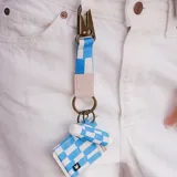 Llavero con cinta de tela a cuadros celestes y blancos, con mosquetón metálico color bronce y aro metálico color bronce.