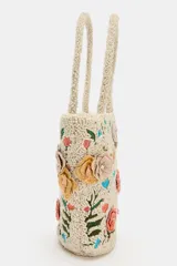 Bolso de mano de formato tipo bucket en color crudo, completamente revestido con abalorios y bordado con motivos florales multicolores en tonos rosados, verdes y celestes. Cuenta con doble asa corta de mano y cierre con botón imantado.