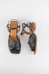 Sandalias de cuero negro con cortes de líneas orgánicas combinados con textil en la capellada, punta cuadrada y taco geométrico de madera. Cuenta con tira ajustable al tobillo con hebilla metálica.