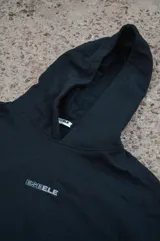 Canguro negro con capucha, corte oversize y diseño con hombros caídos. Presenta detalles bordados en la espalda con la palabra "Dual" en tipografía cursiva junto a texto descriptivo en tonos monocromáticos.