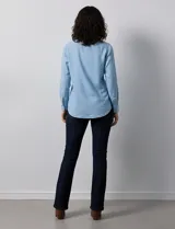 Camisa de manga larga en tela denim de color azul medio, con cuello clásico, botones frontales y dos bolsillos de parche con solapa en el pecho.