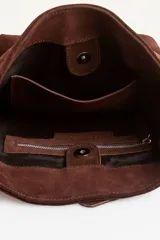 Cartera tipo balde color tostado, confeccionada en gamuza, con correa ajustable decorada con tachas metálicas plateadas.