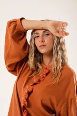 Camisa color naranja óxido con cuello mao, mangas largas abullonadas y cierre frontal con botones. Presenta un volante vertical con borde rojo en el frente.