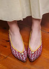 Bailarina de diseño artesanal confeccionada en rejilla bordada con patrón floral en tonos rojizos sobre fondo claro. Presenta ribetes y talón en color amarillo vibrante, con un detalle de media esfera metálica dorada en la puntera y suela plana.