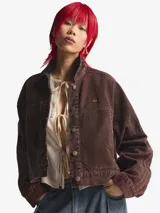 Chaqueta de lona de algodón en color marrón con efecto desgastado, corte holgado, cuello camisero y cierre frontal con botones metálicos. Presenta un pequeño logo de Adidas en tono dorado en el pecho y detalles de costuras decorativas en las mangas.
