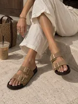 Sandalia plana tipo Birkenstock modelo Lena, con dos tiras anchas en color beige, hebillas metálicas doradas y plantilla de madera oscura.