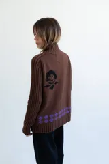 Cardigan de punto color marrón con cierre frontal, cuello alto y bolsillos aplicados. Presenta diseño tejido localizado con detalles en violeta y negro en el pecho.