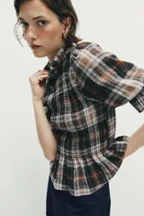 Blusa de cuello subido con diseño de cuadros en tonos marrones, negros y blancos. Presenta mangas cortas con terminación en volantes y un corte ajustado con detalle de volado en la cintura.