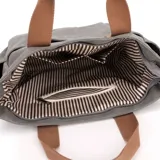 Bolso tipo tote bag de lona color negro, con múltiples bolsillos exteriores y asas cortas y correa larga de tela color marrón. Presenta un parche de cuero marrón con logo grabado en el frente.