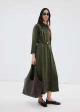 Vestido largo verde militar, con cuello camisero, cierre frontal con botones, mangas tres cuartos y diseño evasé. Incluye un cinturón fino de poliuretano con hebilla metálica.