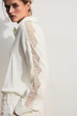 Camisa blanca de manga larga con cuello solapa, cierre frontal con botones y bajo asimétrico. Presenta detalles de puntilla combinada a tono en las mangas y hombros.