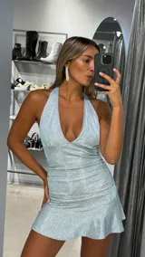 Vestido corto ajustado, color celeste claro con acabado brillante (lurex o glitter). Presenta un escote halter profundo en V y frunces laterales que acentúan la silueta. La falda tiene un ruedo con volado.