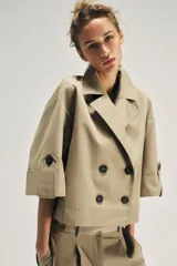 Trench corto de color beige, con cuello de solapa, cierre cruzado con botones y mangas cortas con vuelta y trabilla con botón.