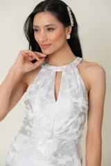 Vestido largo de fiesta en gasa labrada color blanco con detalles plateados, con cuello halter, drapeado en la cintura y tajo en la parte trasera.
