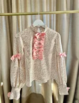Blusa de cuello alto con cierre de botones en la espalda, decorada con una hilera vertical de lazos rosados en el frente. Presenta mangas largas abullonadas con puños ajustados y lazos decorativos en los codos. Confeccionada en una tela con estampado floral de pequeñas flores rosas y verdes sobre fondo claro.