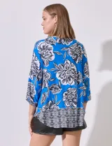 Blusa de crepe estampada con flores blancas y negras sobre fondo azul. Tiene escote entrelazado con abertura, mangas 3/4 con puños elastizados y corte holgado.