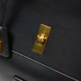 Cartera tipo satchel negra de material sintético (PU) con forrería textil. Presenta un compartimento principal amplio con cierre de solapa y herraje metálico dorado. Incluye bolsillo interno con cierre, correa larga regulable y desmontable, y logo XL aplicado en el frente.