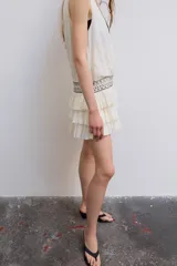 Vestido corto de gasa color blanco, con escote en V cruzado, sin mangas y diseño con volantes en el bajo. Presenta un detalle de tachas plateadas en la cintura.