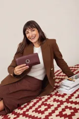 Billetera larga de símil cuero color marrón claro, con solapa frontal decorada con un aro metálico central. Cuenta con múltiples compartimentos internos para tarjetas, billetes y un monedero con cierre.