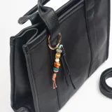 Cartera tipo tote de estructura rígida y diseño minimalista en color negro. Presenta doble asa de mano y una correa larga ajustable y extraíble. Incluye un detalle decorativo de argolla metálica con cuentas de colores y tiras de cuero sintético colgantes.