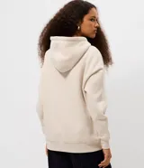 Buzo canguro de corte oversized color beige, con capucha ajustable mediante cordones y bolsillo frontal. Presenta una estampa estilo college en el pecho con la inscripción 'West Coast 1994' en color azul marino.