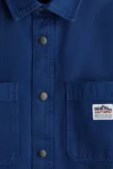 Sobrecamisa azul de lona gruesa de algodón, con cuello, tapeta con botones a presión y canesú en la espalda. Tiene mangas largas con botón a presión en los puños y dos bolsillos superiores de parche. El bajo es levemente redondeado y el corte es oversized.