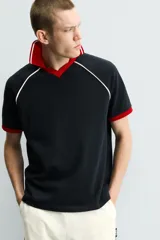 Polo de corte relajado confeccionado en tejido de algodón con estructura abierta. Presenta cuello polo con abertura frontal en V, mangas cortas tipo ranglán y acabados en rib a contraste en color rojo y blanco.