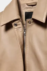 Blazer de cuero color beige con cuello solapa, manga larga y cierre frontal con cremallera y botones a presión ocultos. Presenta bolsillos laterales ocultos en costura y forro interior.