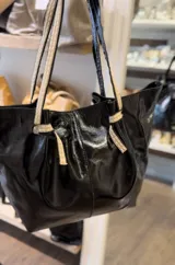 Bolso tipo tote de cuero negro con acabado brillante, diseño fruncido en los laterales y correas de hombro de tela tejida en tono beige.