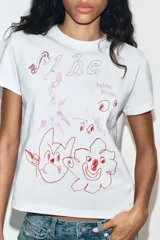 Camiseta de algodón blanca con cuello redondo y manga corta. Presenta un estampado gráfico frontal con dibujos estilo garabato y texto en tonos rojizos.