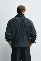 Jersey de punto grueso con estructura de volumen, corte relajado, cuello redondo y manga larga con puños y ruedo acanalados. Presenta un diseño bicolor con trama en tonos verde oscuro y azul.