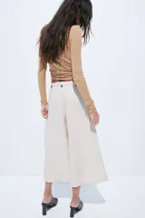 Pantalón sastrero cropped de tiro alto, color beige, con piernas amplias, pliegues frontales, bolsillos al bies y trabillas en la cintura con detalles de botones.