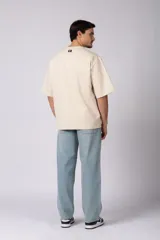 Remera de corte oversize color beige, con cuello redondo y etiqueta decorativa pequeña en el ruedo inferior.