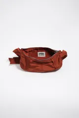 Bolso tipo hobo de la marca Bimba y Lola, modelo Moon mediano, de color rojo óxido. Confeccionado en nylon con diseño de media luna, cierre superior con cremallera, correa ajustable con hebilla metálica y el logotipo de la marca impreso en la parte frontal.