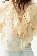 Blusa de gasa arrugada de corte holgado, en tono amarillo claro con estampado floral. Cuenta con escote en V profundo, un amplio volante ribeteado con encaje alrededor del cuello y cordones finos para atar al frente. Mangas largas con puños elásticos.