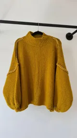 Buzo tejido de punto color mostaza, con cuello alto, diseño oversize y mangas abullonadas. Presenta detalles de costuras decorativas bordadas a mano en color blanco en las uniones de las mangas.
