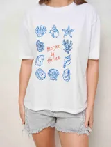 Remerón de algodón color blanco, de corte oversize, manga corta y cuello redondo. Presenta un estampado central con ilustraciones de caracoles y conchas marinas en azul, junto con el texto "Meet me by the sea" en tono rojizo.