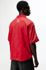 Camiseta de futbol de manga corta de la marca Lotto, en color rojo con un sutil estampado geométrico. Cuenta con cuello tipo polo abierto con escote en V y un calce holgado. Las mangas presentan una franja blanca lateral decorada con el logo de la marca repetido. El logotipo de Lotto destaca en blanco en el centro del pecho.