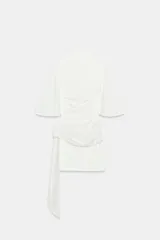 Vestido corto blanco con mangas cortas amplias y cuello recto. Presenta un fajín drapeado de lentejuelas plateadas a la cintura con un lazo largo lateral.