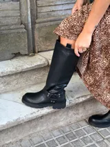 Bota de caña alta estilo biker, confeccionada en cuero sintético negro. Presenta una suela track con plataforma, puntera redondeada y un detalle de arnés con hebilla metálica y tachas en la zona del tobillo.