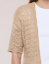 Bolero corto de punto beige, con diseño abierto sin botones y mangas cortas. Presenta un patrón de tejido calado con detalles geométricos y terminaciones onduladas en los bordes.