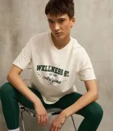 Remera de algodón color crema, de corte amplio y mangas cortas. Presenta cuello en V y estampa frontal con texto en verde y negro.
