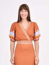 Blusa color ladrillo con escote cruzado, mangas cortas abullonadas con detalle de estampado de ondas en lila y naranja, y ribete de conchillas en escote, cintura y mangas.