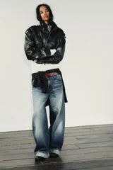 Cazadora corta de efecto piel color marrón oscuro, con cuello subido, manga larga con trabillas en los hombros, bolsillos con solapa en el delantero y cierre frontal con cremallera y botones a presión ocultos por solapa.