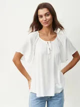 Blusa blanca de manga corta con textura arrugada, escote redondo con abertura en V y lazos ajustables. Presenta detalles de puntilla calada en los hombros.