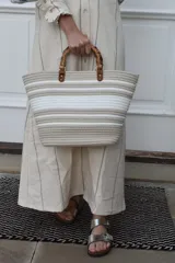 Bolso tipo tote color beige con rayas horizontales blancas, asas de mano de bambú y detalles en cuero sintético marrón con herrajes dorados.