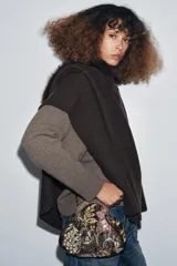 Bolso pequeño tipo sobre con cierre de clip metálico, confeccionado en tela marrón con bordados florales multicolores y aplicaciones de abalorios. Incluye una cadena metálica fina para llevar al hombro.